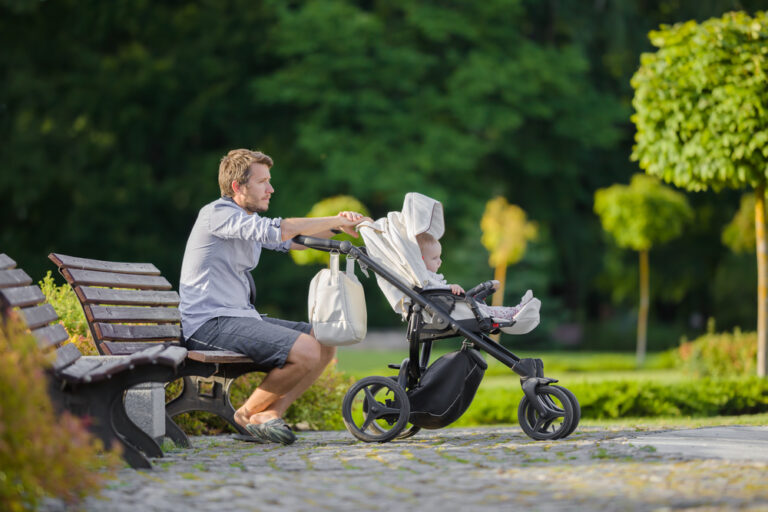 Young,Man,Sitting,On,Wooden,Bench,At,Town,Green,Park